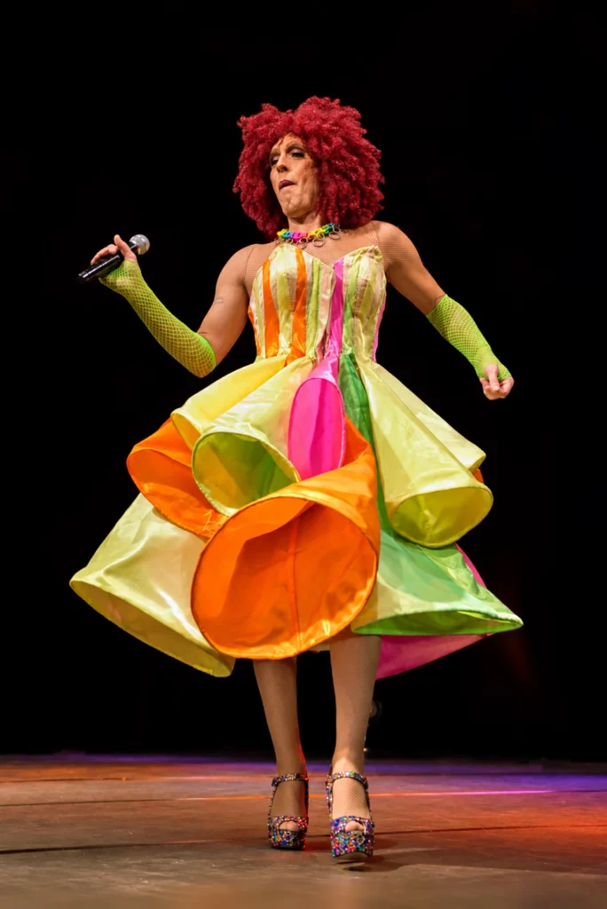 Miss Caline sur scène en robe multicolore à grands volants de satin, perruque rousse frisée et mitaines vertes, tenant un microphone.