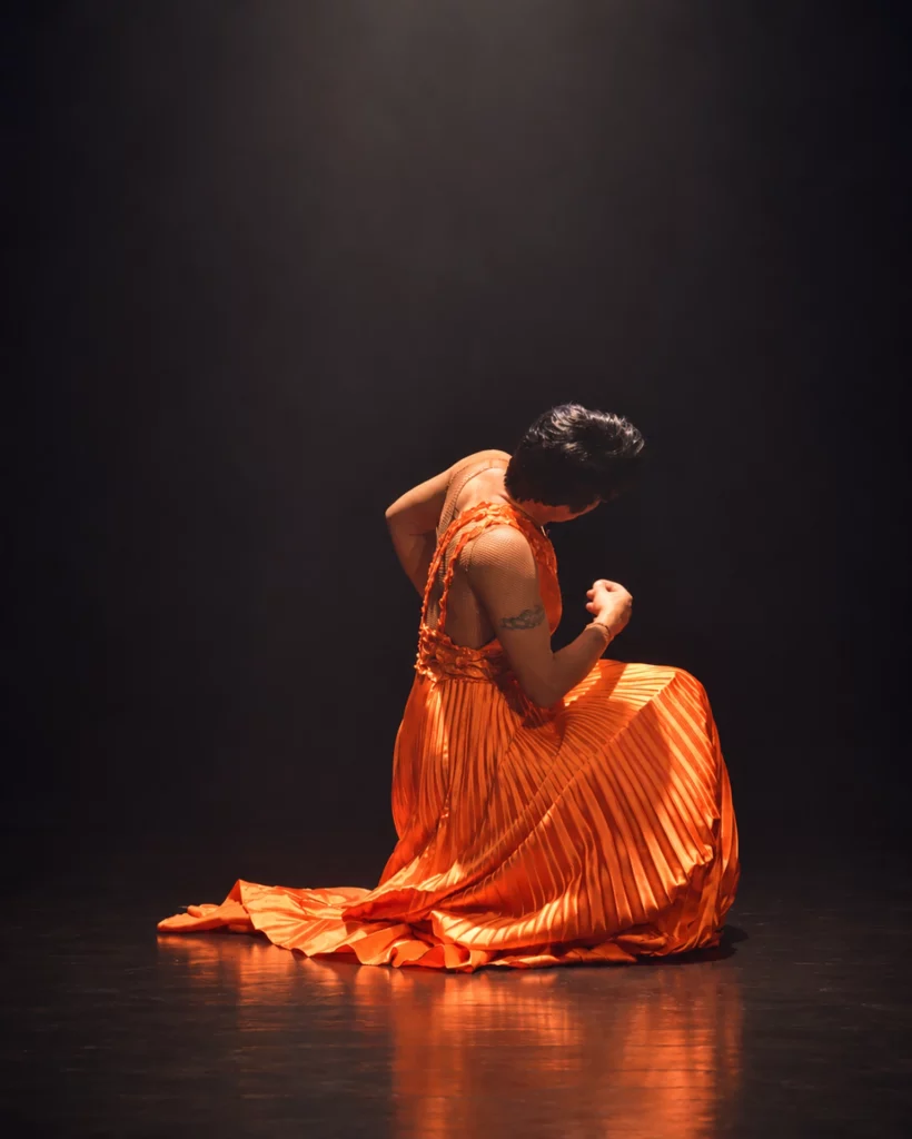 Miss Caline agenouillée sur scène dans une longue robe orange plissée, sous un éclairage tamisé, sur fond noir.