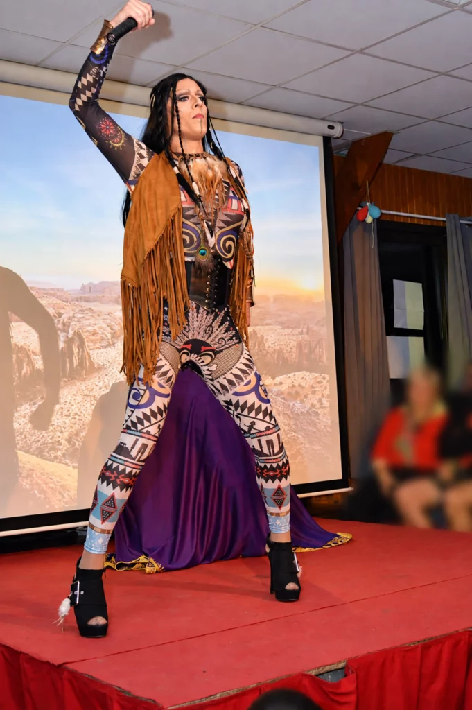 Miss Caline sur scène, avec un maquillage féminin prononcé, une longue perruque noire et un microphone levé.