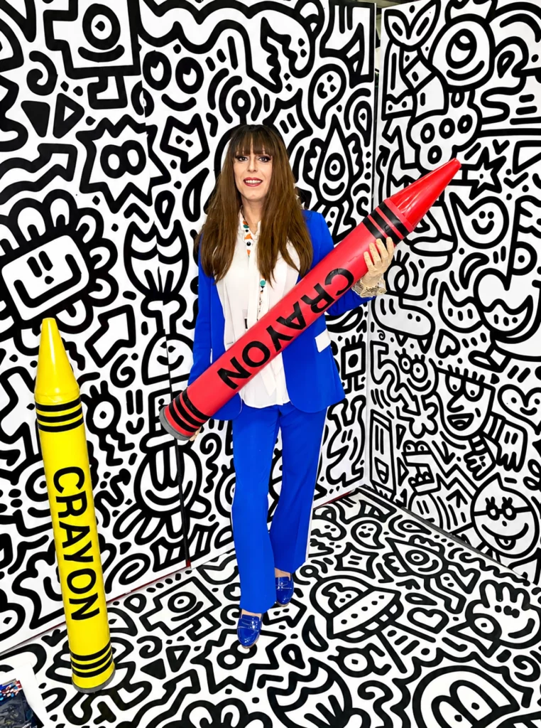 Miss Caline en tailleur bleu électrique, tenant un crayon géant rouge, dans un décor graphique noir et blanc, à côté d'un crayon jaune.
