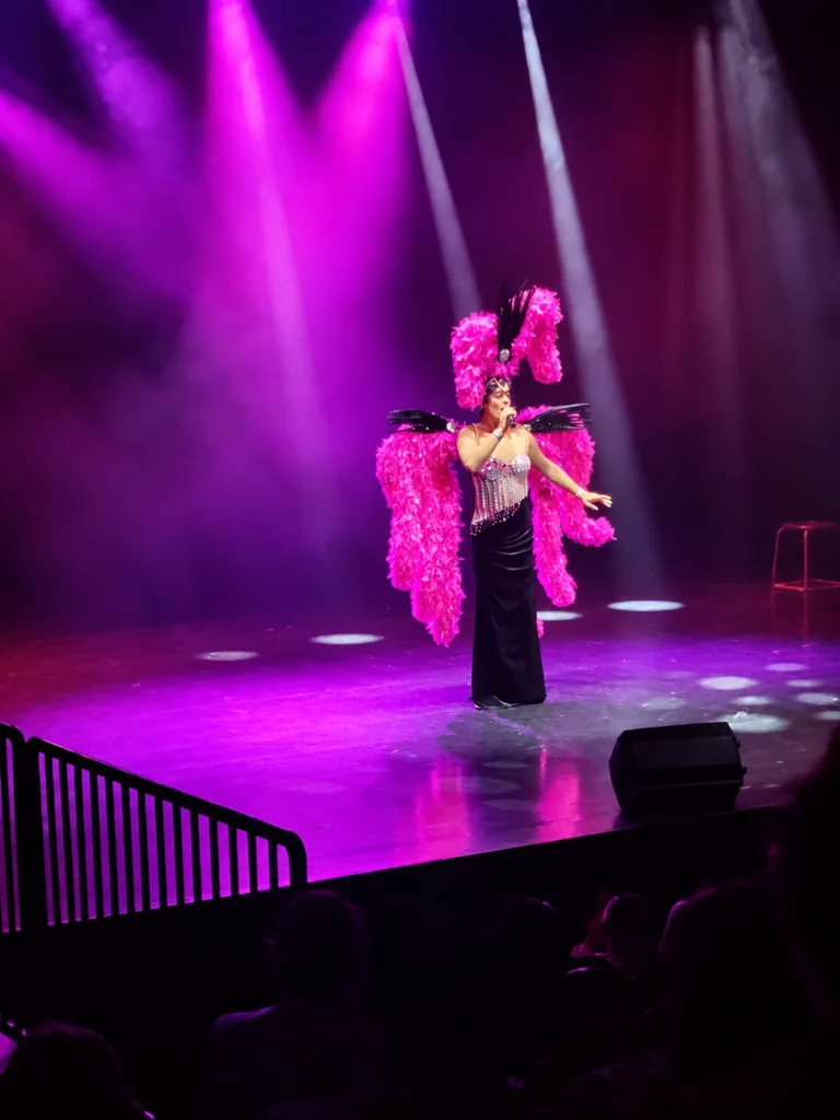 Miss Caline seule sur scène, en longue robe noire et costume à grandes plumes roses, sous des projecteurs violets, un microphone à la main.