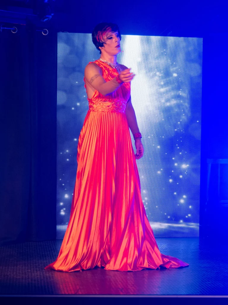 Miss Caline sur scène dans une longue robe orange lors de son spectacle transformiste