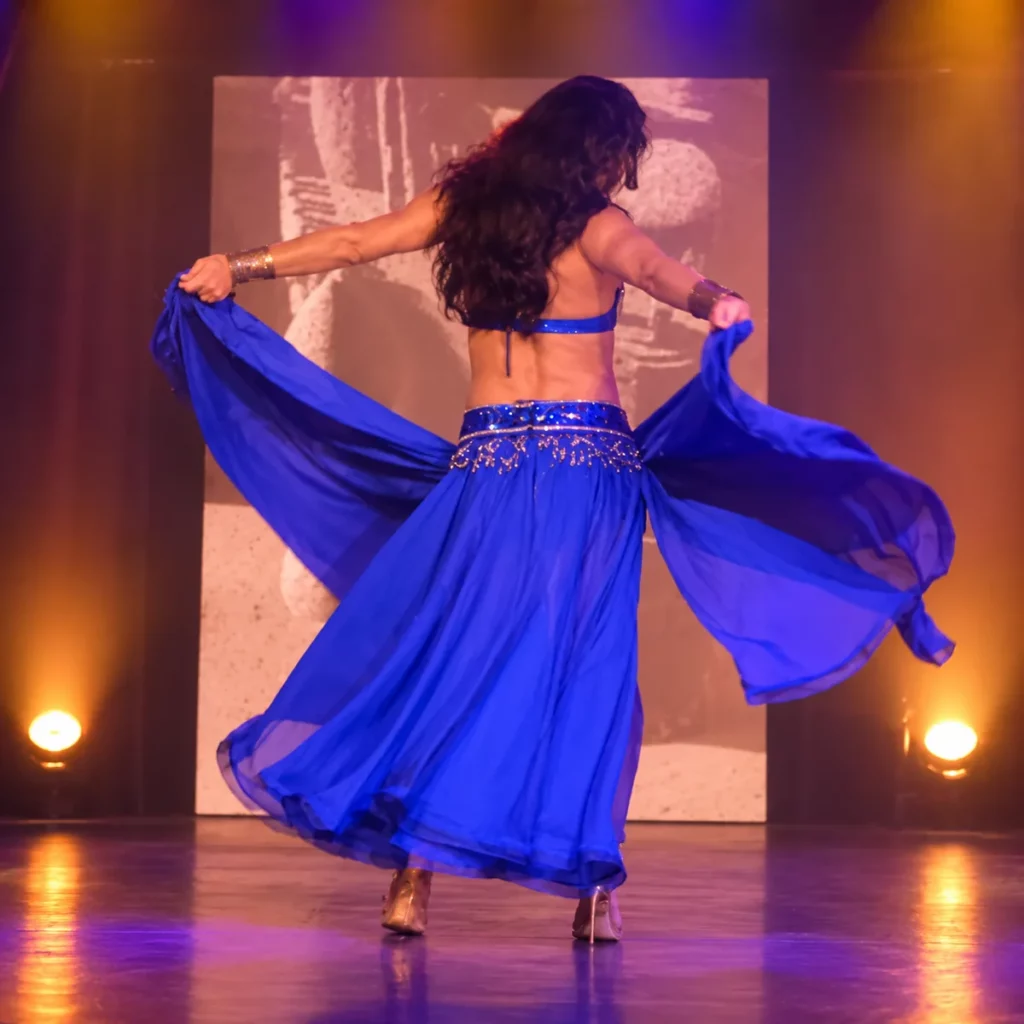 Miss Caline, transformiste, vue de dos sur scène dans un numéro de danse orientale, longue chevelure brune, costume bleu à sequins et jupe en voile déployée à deux mains